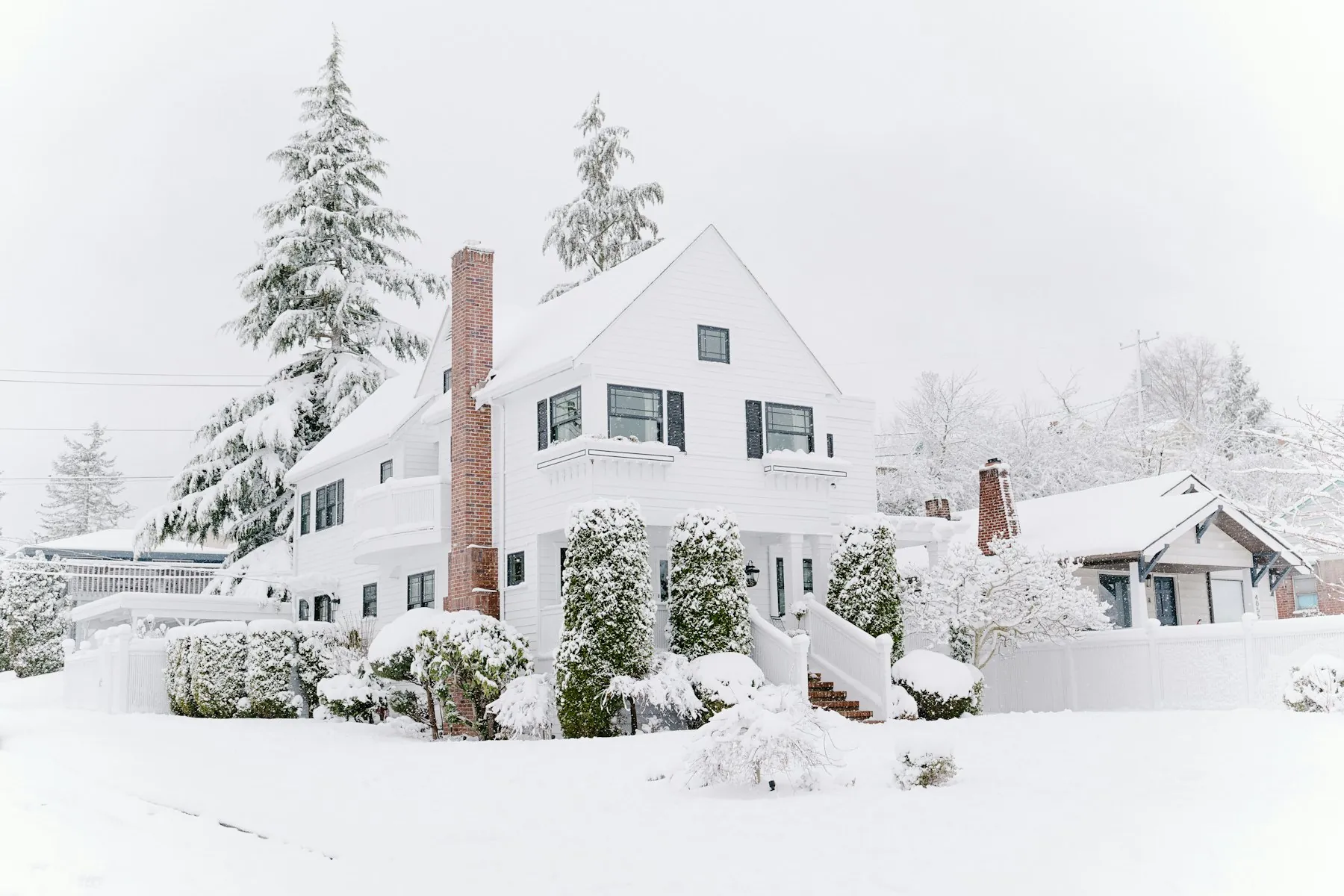 A white house with a brick chimney surrounded by fresh snow and evergreens